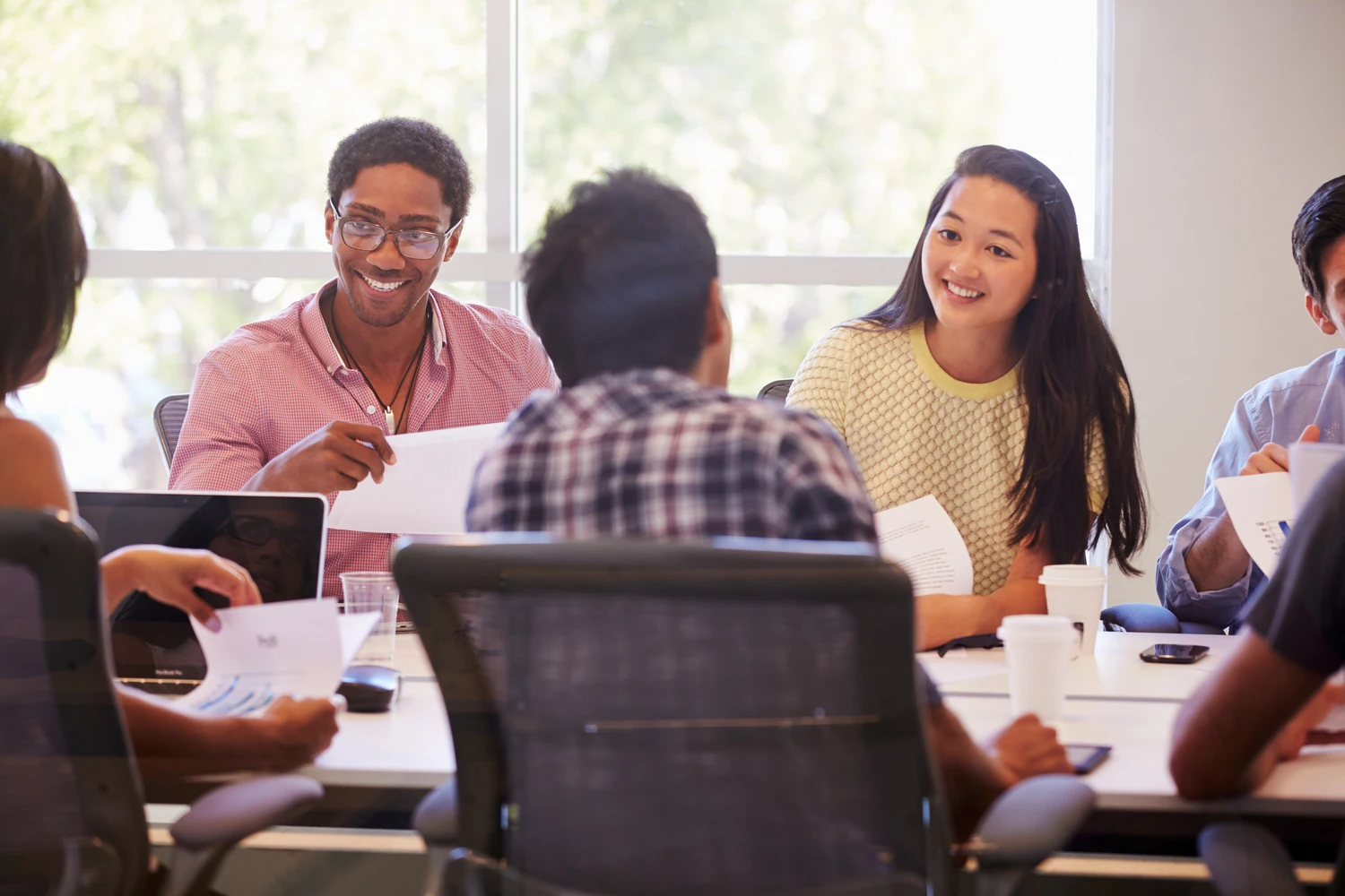 A diverse group of people engaged in a meeting, discussing documents around a conference table with laptops and coffee cups.
