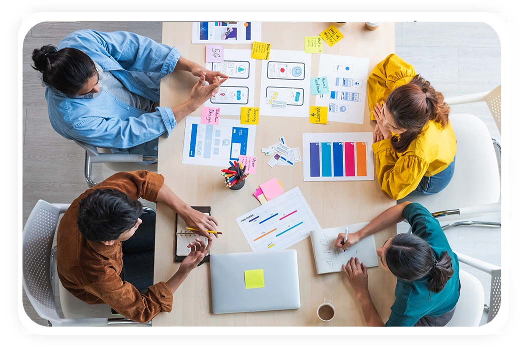 A diverse group of individuals collaborates around a table filled with design documents, colorful markers, and a laptop.