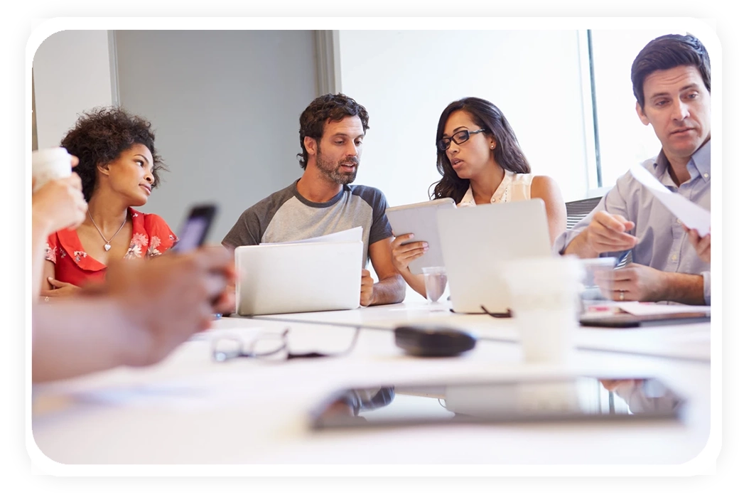 A diverse group of people engaged in a meeting, discussing ideas with laptops and papers on the table in a bright conference room.